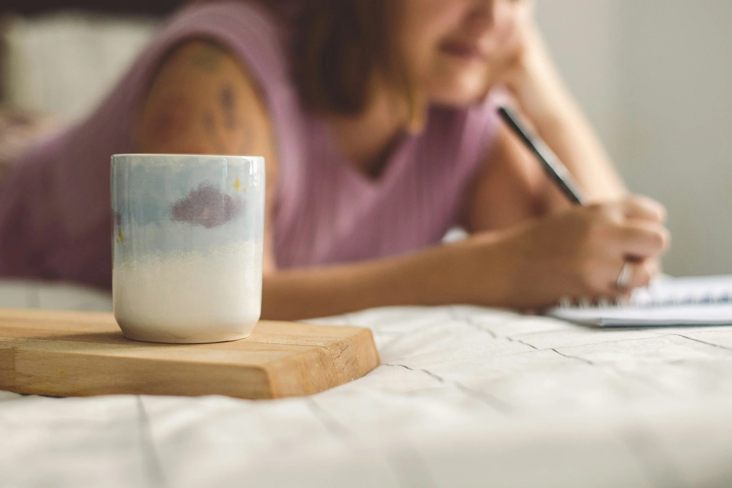 Coffee mug on a bedside table while a woman writes in a notebook on the bed