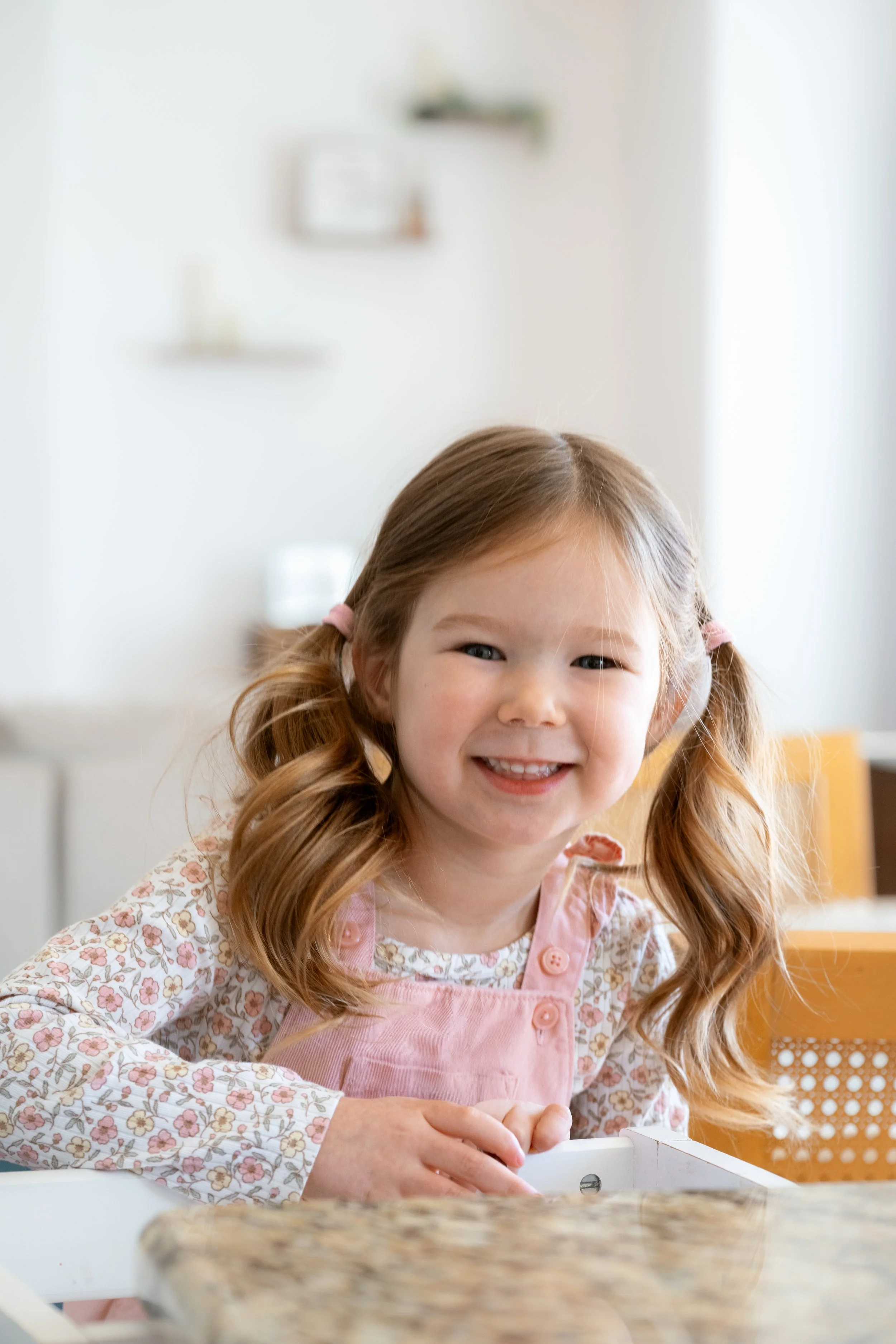 A smiling young girl with pigtails, wearing a pink apron, sitting at a kitchen table.