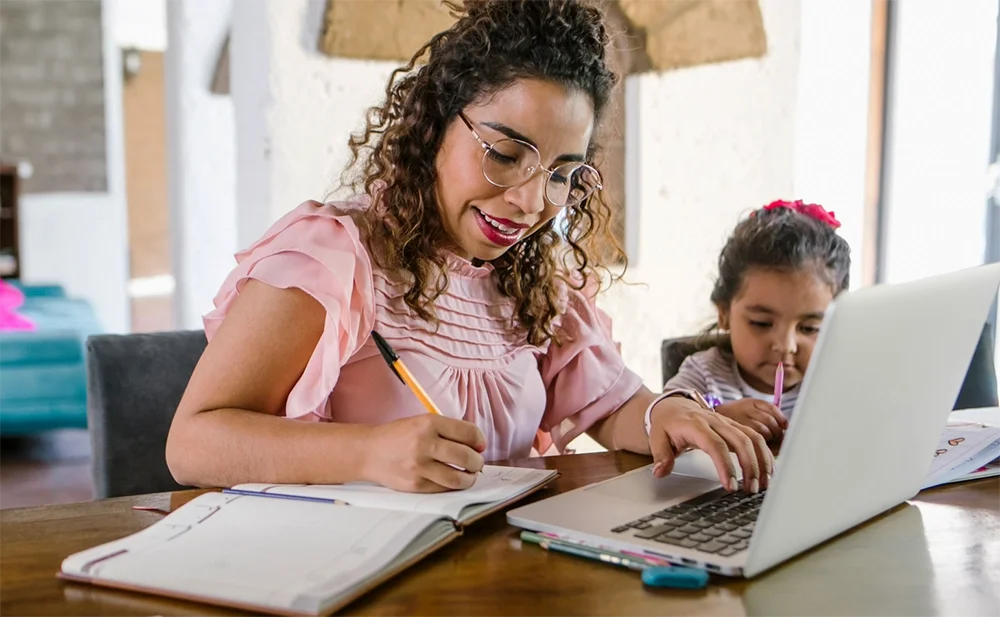 A smiling woman with glasses is writing in a notebook and typing on a laptop, working from home while a young child sits next to her.