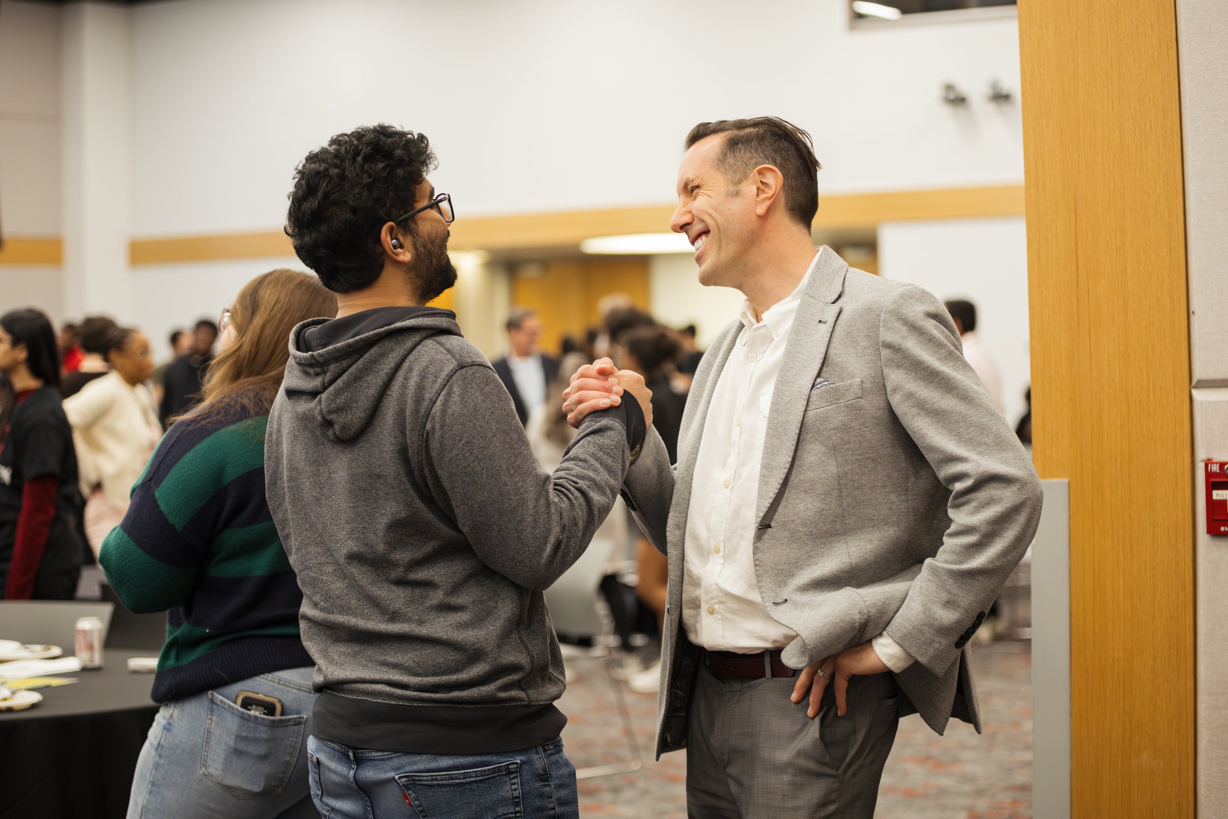 Two men—one in a grey hoodie and the other in a grey suit jacket—are smiling and shaking hands vigorously in a professional networking or conference setting.