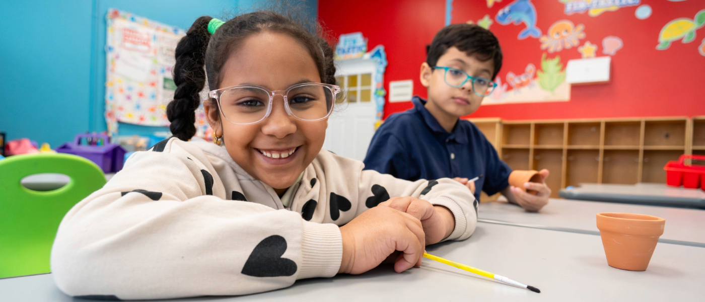 Two children sitting at a classroom table with a small terracotta pot in front of them. The girl in the foreground is smiling and has glasses, while the boy in the background is looking at his phone.