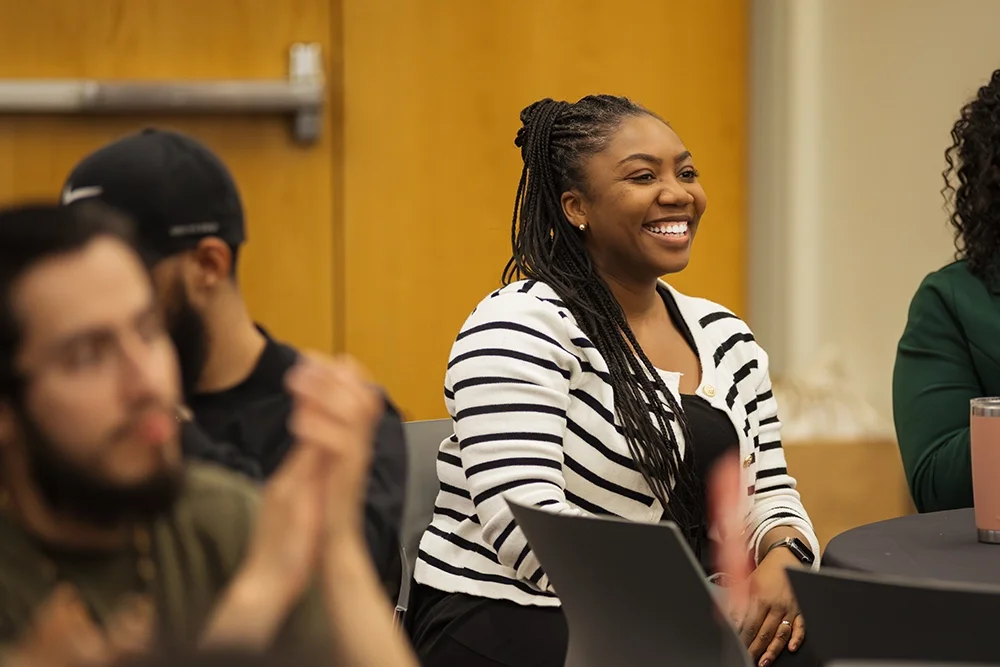 A smiling woman with braids, wearing a black and white striped cardigan, is sitting and looking toward an unseen speaker at a public event.