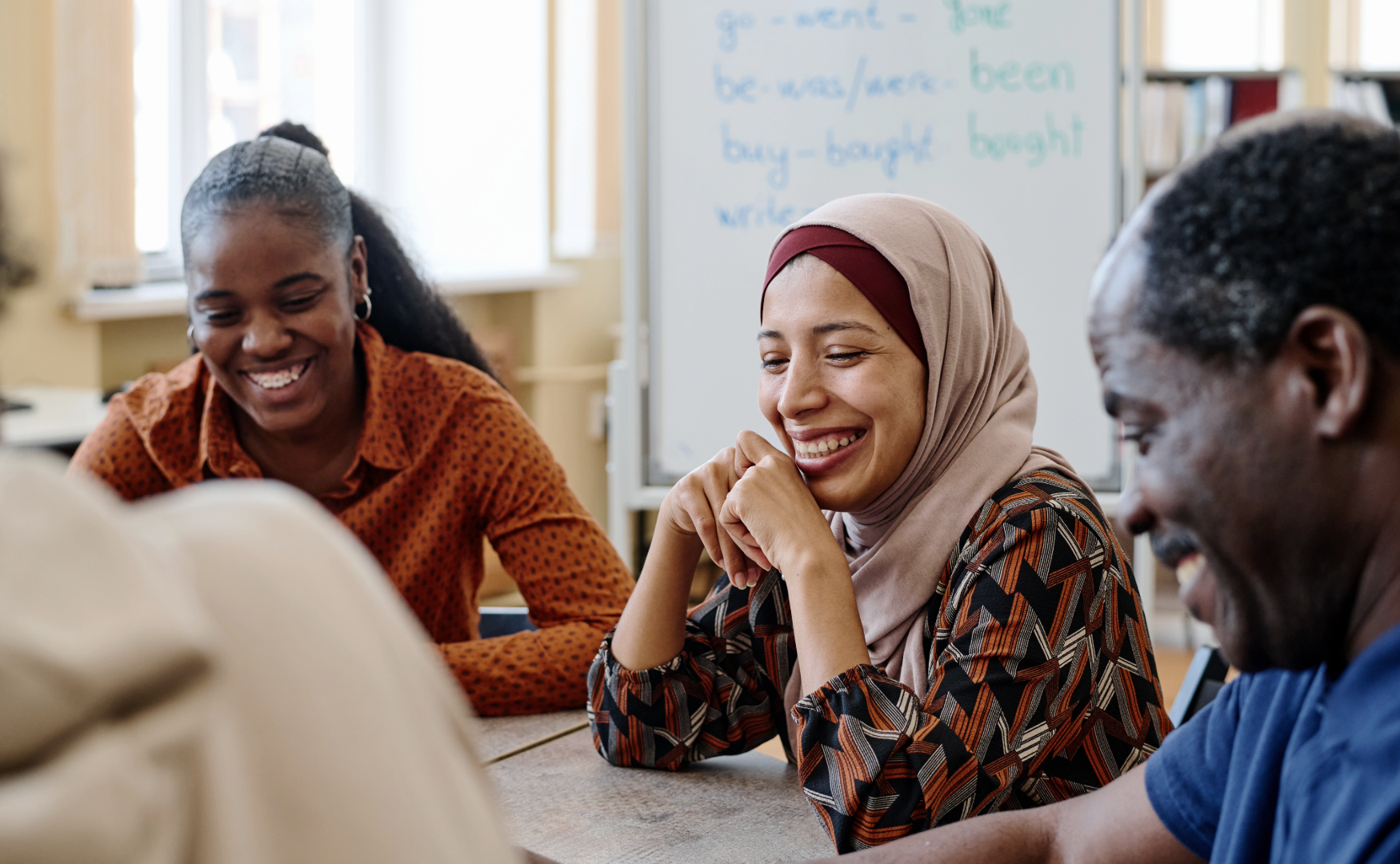 A group of diverse adults smiling and laughing during a discussion in a classroom or meeting room with a whiteboard in the background.