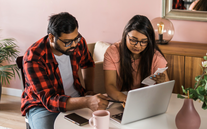A man and a woman sitting at a table working on a laptop, with a receipt and a phone in front of them, in a cozy room with pink walls, plants, and decorative items.