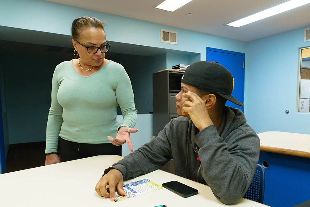 A woman in glasses and a light green sweater is speaking to a young man wearing a black cap and gray hoodie, sitting at a table with a phone, a flyer, and a pen in front of him in a room with blue walls.
