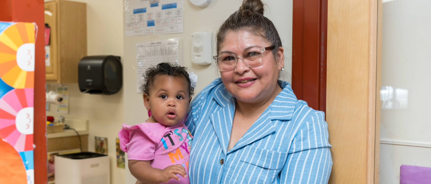 A woman with glasses smiling, holding a young girl in a pink shirt, in a room with colorful decorations and educational materials.