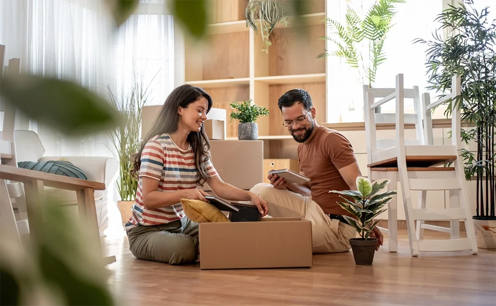A smiling couple is sitting on the floor in their new, sparsely furnished home, unpacking a cardboard box near a small potted plant.