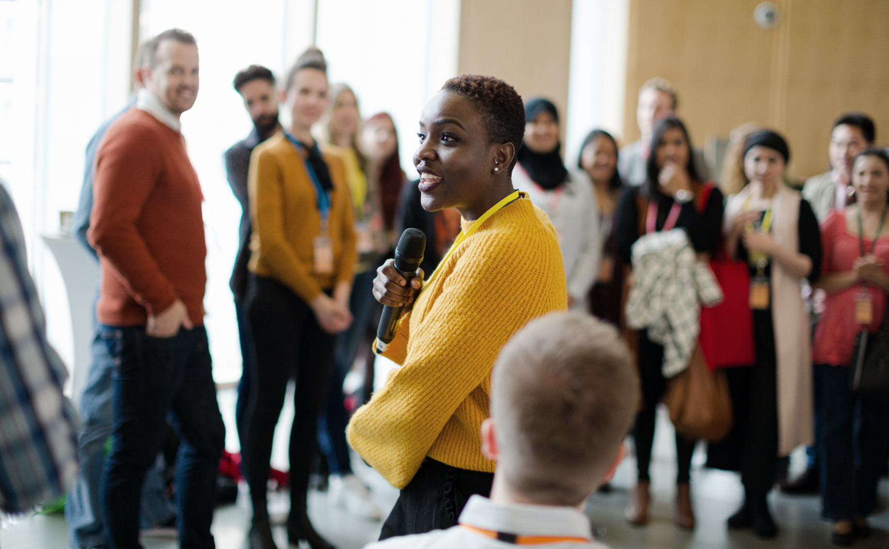 A woman in a yellow sweater holds a microphone and speaks to a group of diverse people at a conference.