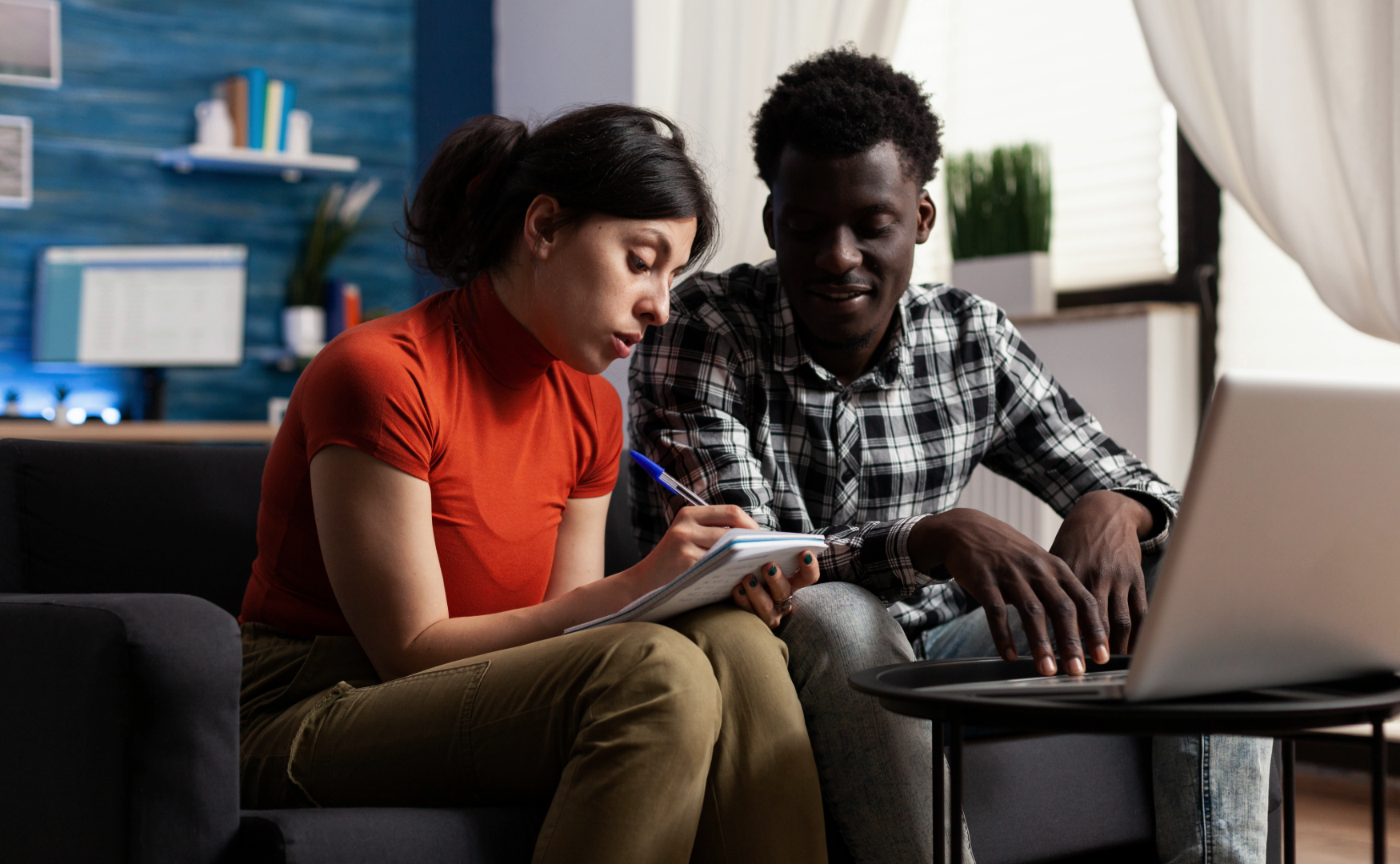 A woman and a man sitting on a black couch, discussing work with a notebook and laptop in front of them in a cozy room.