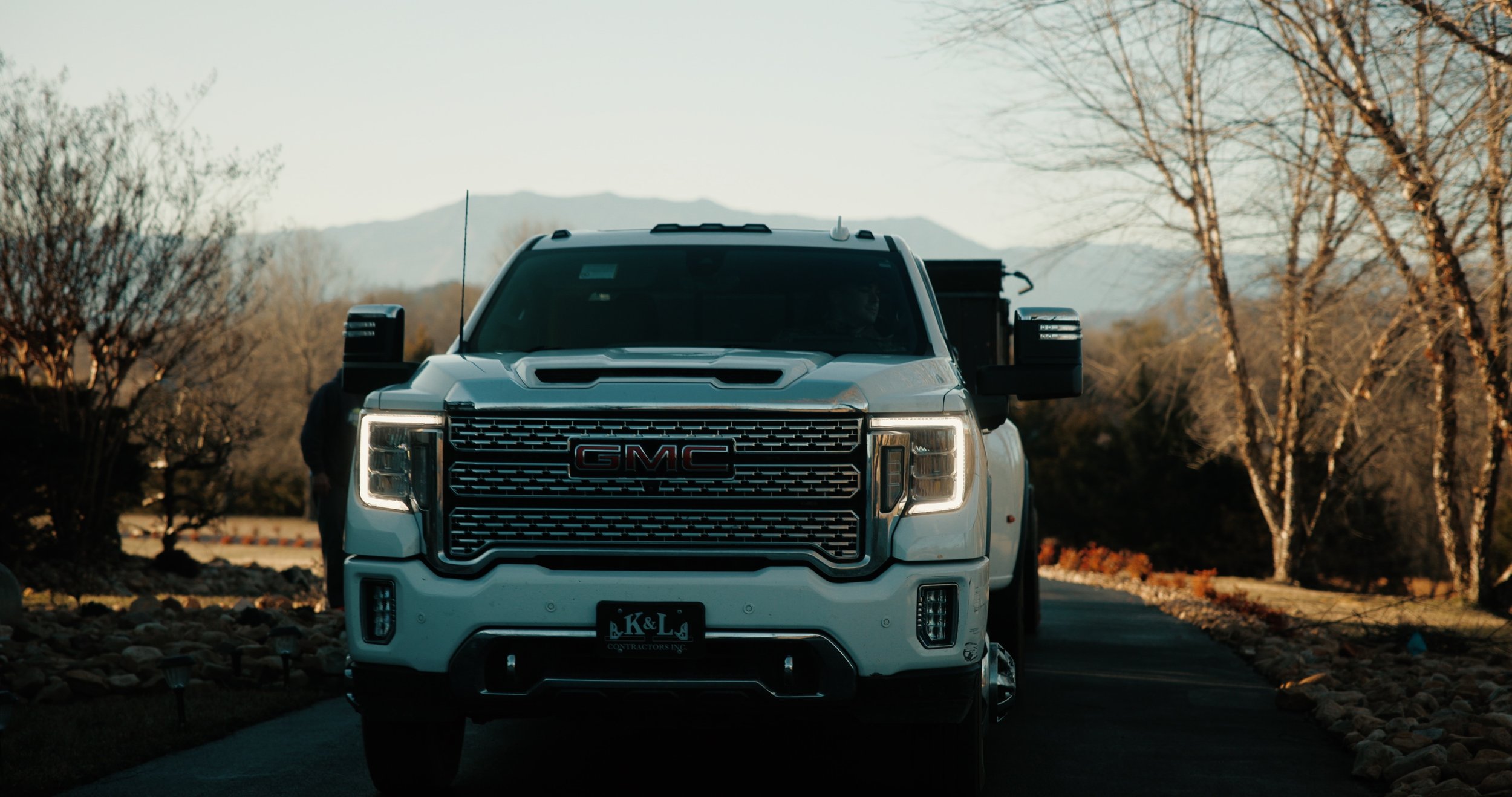 A white GMC truck driving down a paved road with trees and mountains in the background.
