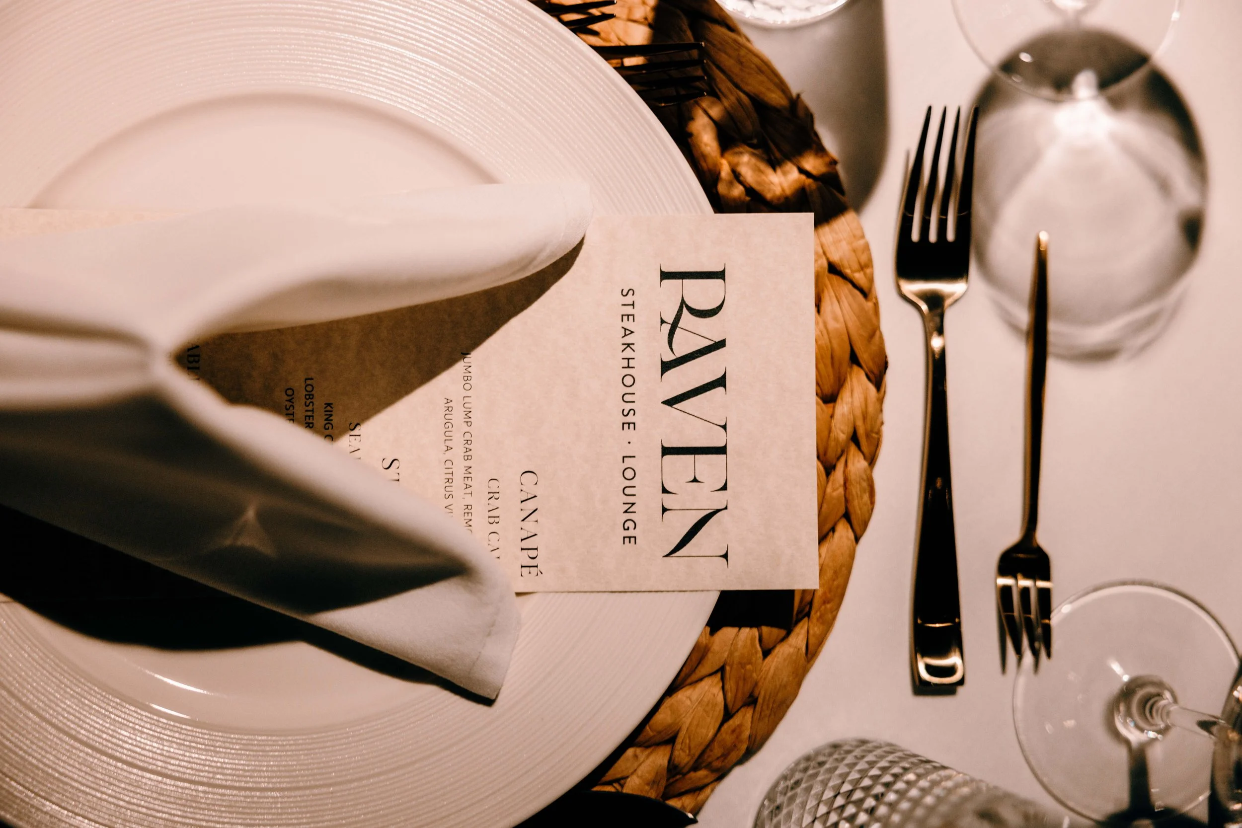 Elegant table setting with a white plate, a folded white napkin, a wine glass, a fork, a knife, and a menu from Ravene Lounge on a woven placemat.