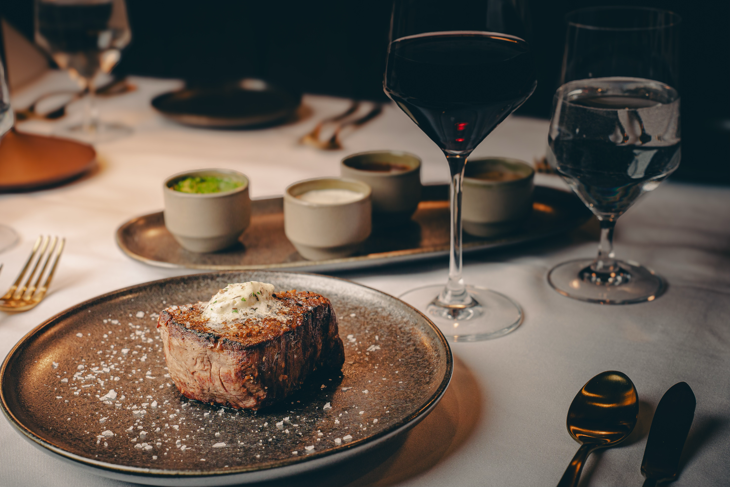 A plated filet mignon steak topped with a dollop of herb butter, served on a dark ceramic plate with coarse sea salt, alongside a glass of red wine, and a wine glass of water, on a white tablecloth with gold flatware, in an elegant dining setting.