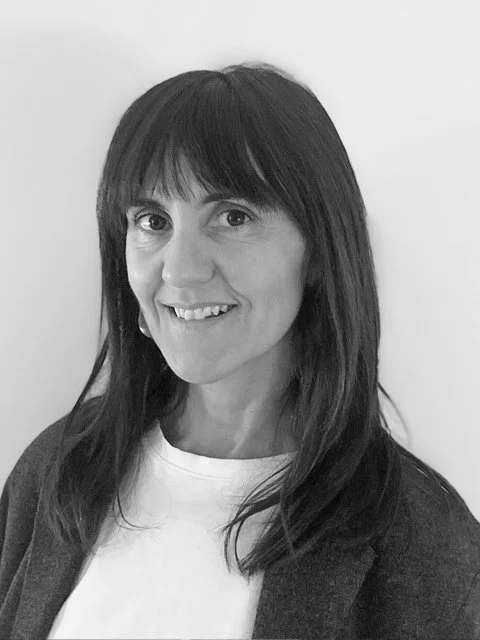 Black-and-white portrait of a woman with long dark hair, smiling, wearing a white shirt and dark jacket, against a plain background.