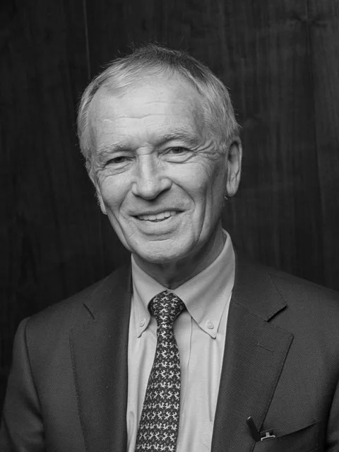 Black and white photo of a smiling older man in a suit with a patterned tie, standing in front of a dark wooden background.