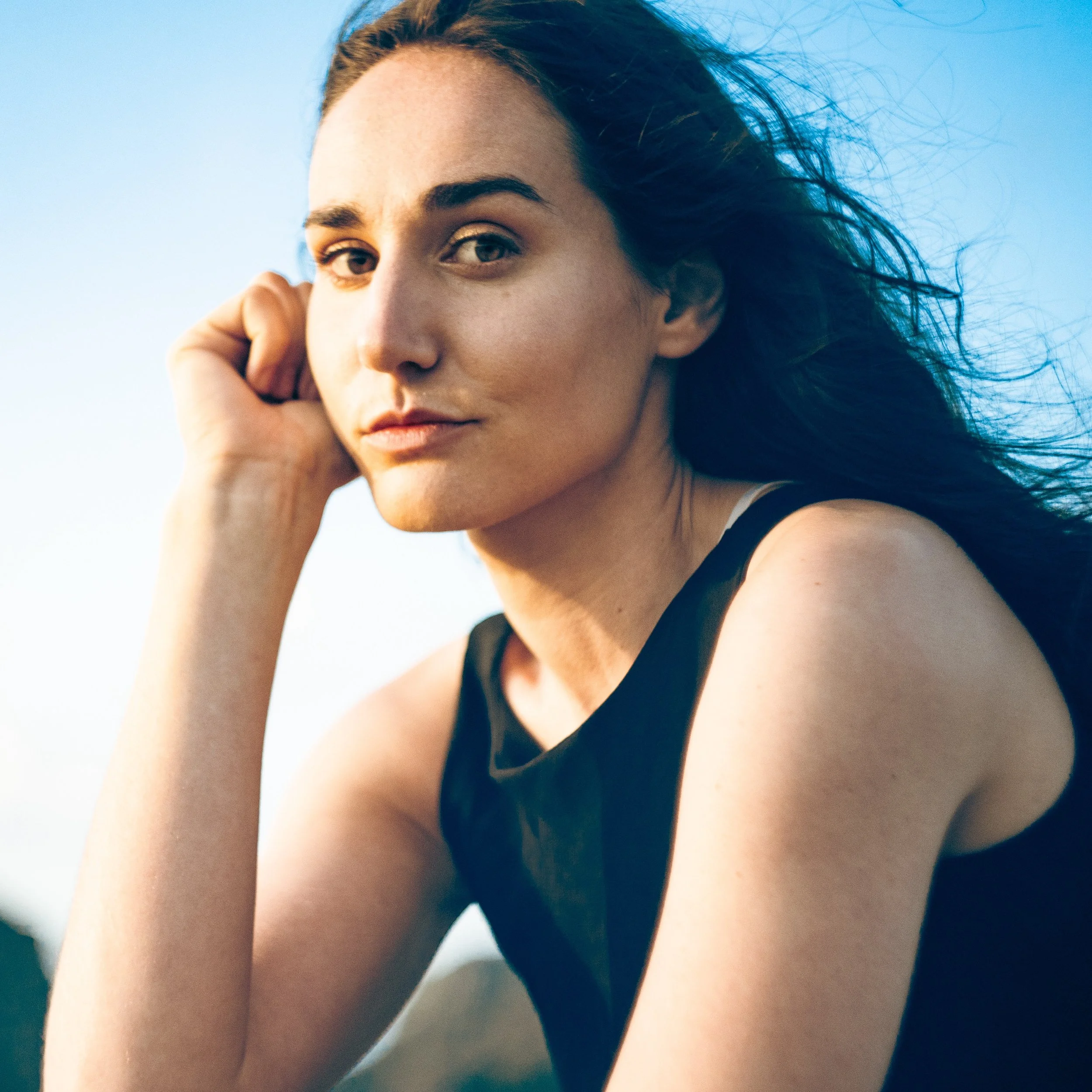 Close-up of a woman with long dark hair, wearing a black sleeveless top, outdoors against a blue sky, with natural sunlight highlighting her face.