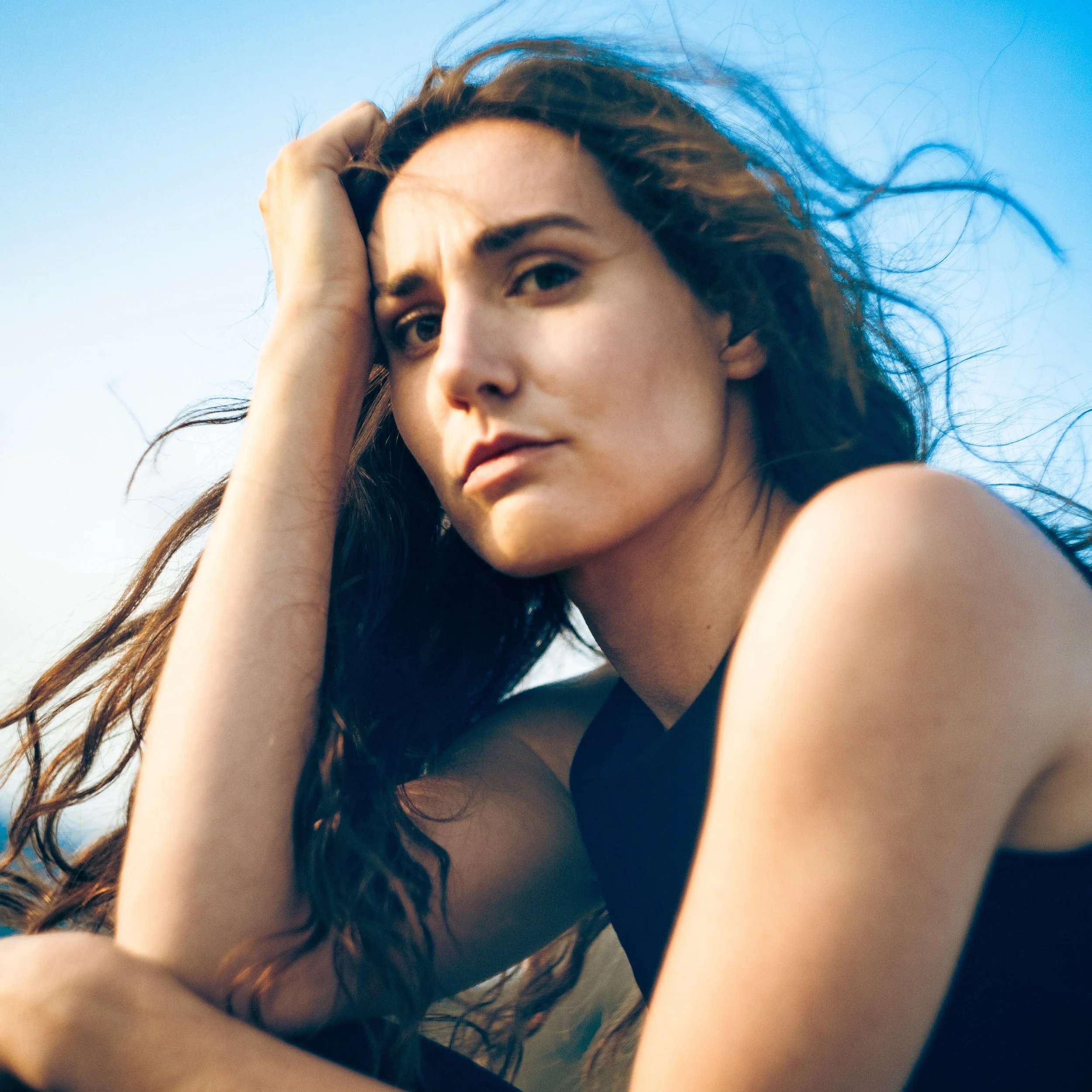 A woman with long dark hair looking into the camera with a serious expression, outdoors against a clear blue sky.