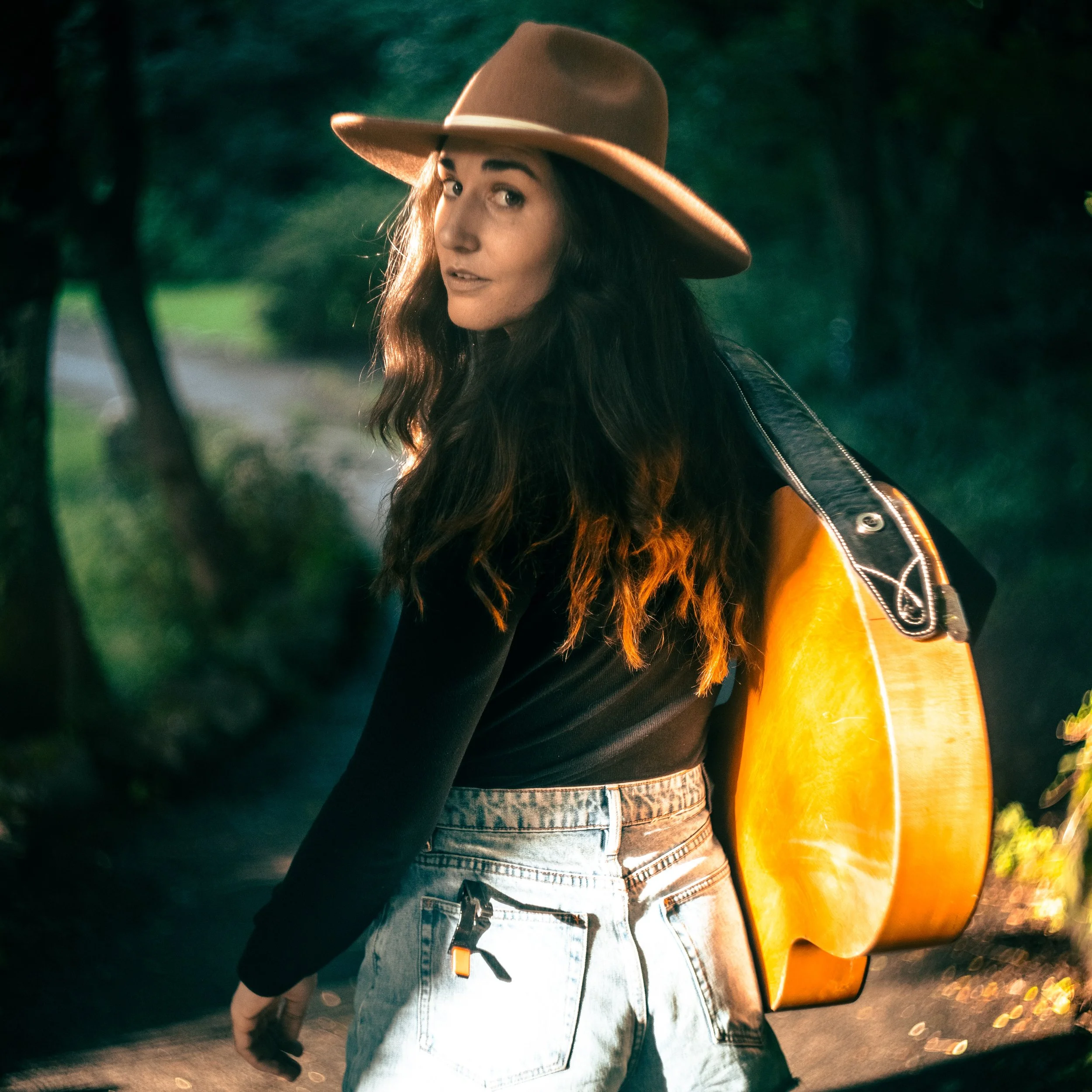 A woman with long, wavy dark hair wearing a wide-brimmed tan hat, black long sleeve top, and light blue jeans, carrying an acoustic guitar on her back, standing outdoors on a path surrounded by greenery.