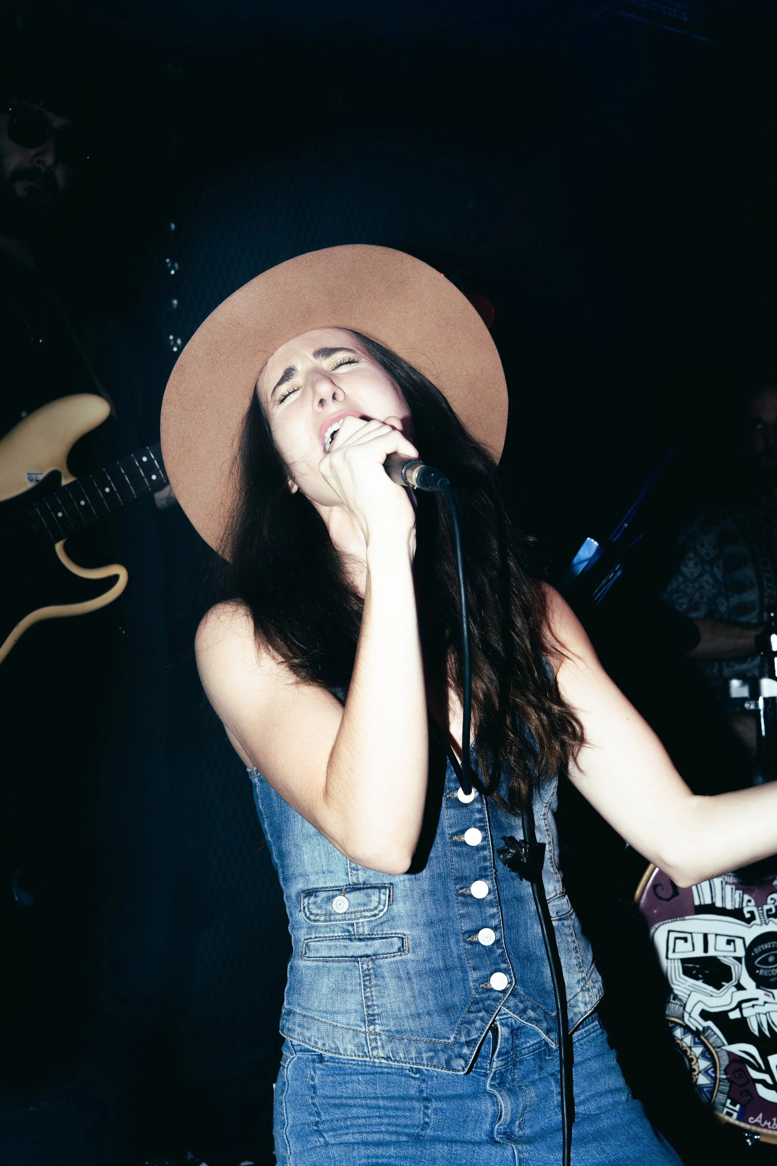 Female singer wearing a wide-brimmed hat and denim outfit singing passionately into a microphone during a performance. A male guitarist is partially visible in the background.