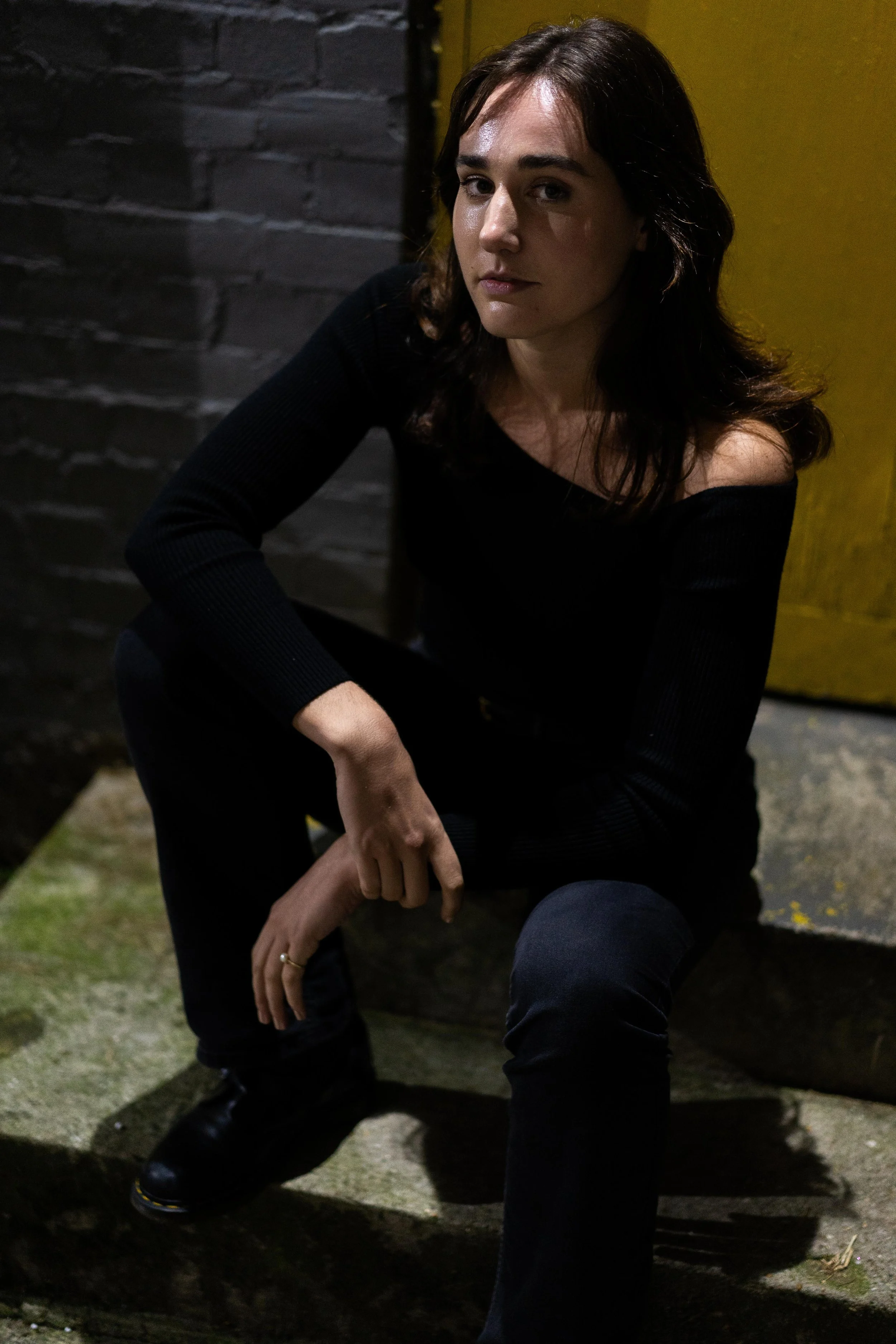 A young woman with dark brown hair and light skin, wearing a black off-the-shoulder sweater, sits on a stone step in front of a brick wall and yellow door, looking at the camera.