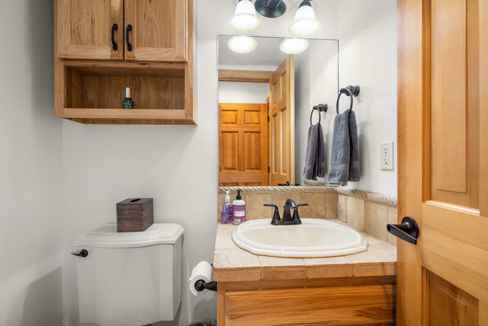 Bathroom with wooden cabinets, a mirror, a sink with black fixtures, a toilet with a tissue box on top, and two gray towels hanging on black towel rings.