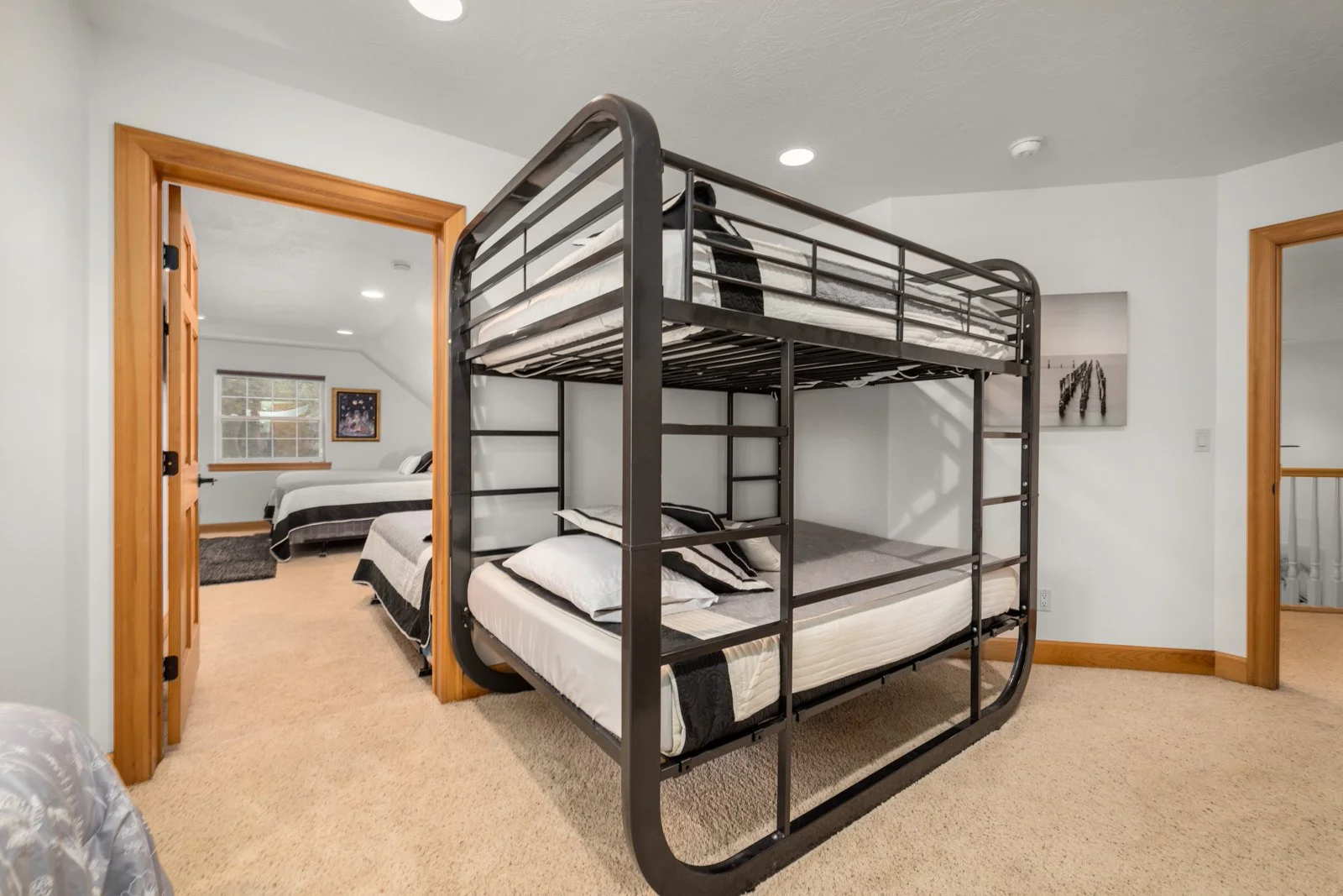 A room with a black metal bunk bed, two beds with white and black bedding. Two additional beds are visible in the adjacent room through an open doorway, each with similar bedding. The rooms have beige carpeted floors and wooden trim around the doorwa