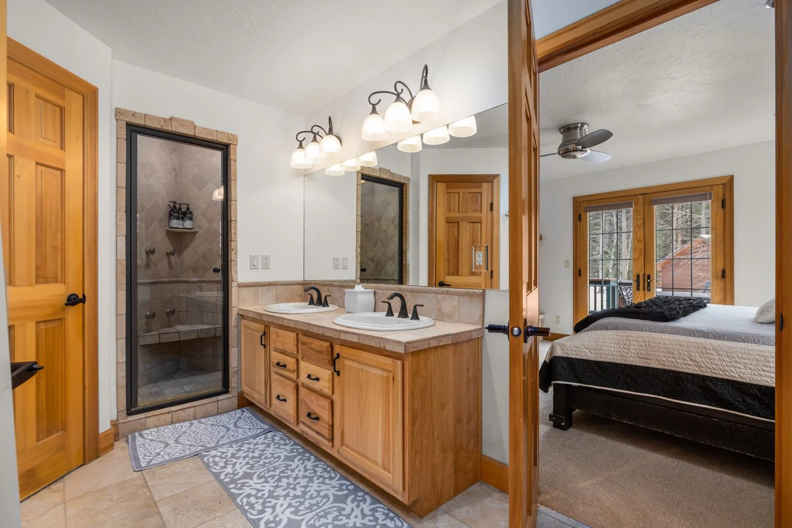 Bathroom with double sinks, wooden cabinets, and a glass shower, connecting to a bedroom with a bed, windows, and a ceiling fan.