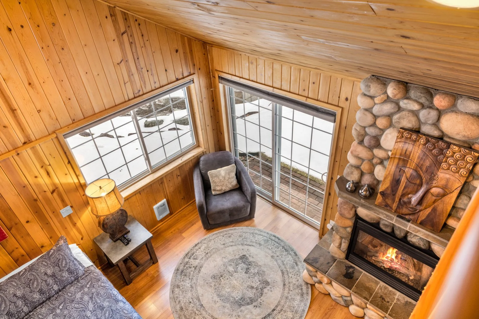 Living room with wood-paneled walls, a stone fireplace with a wooden carved art piece above, a large window, and a sliding glass door, with a gray armchair, a round rug, and a side table with a lamp.