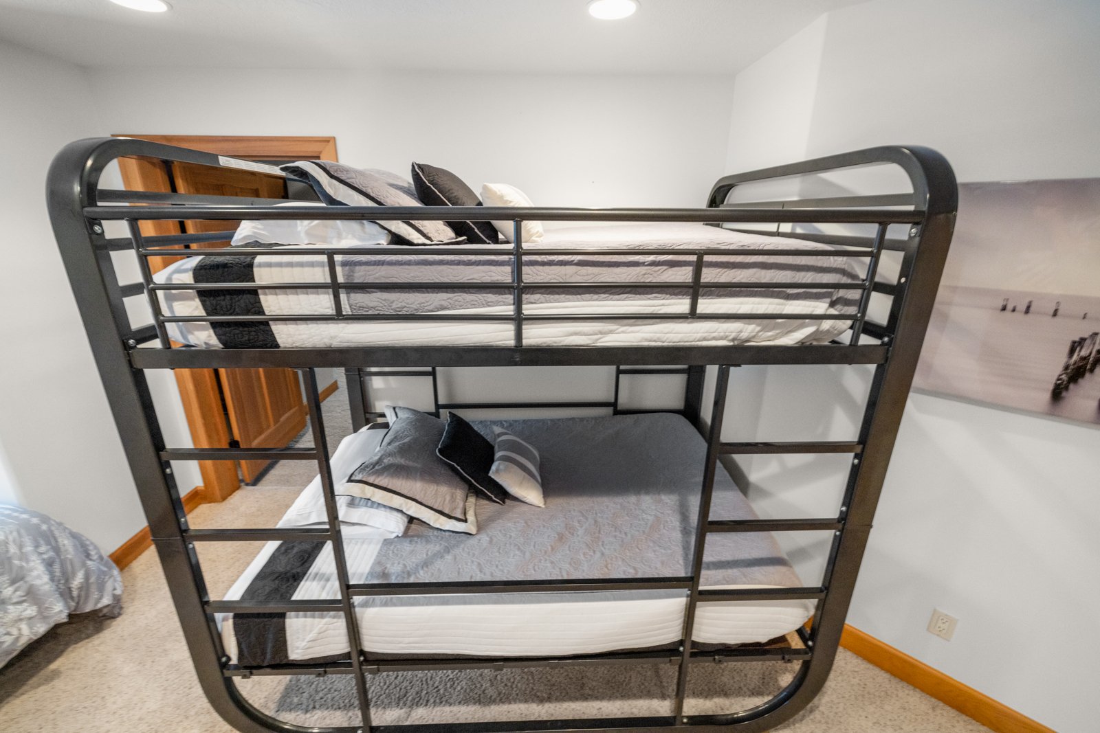 A black metal bunk bed with two mattresses, pillows, and bedding, situated in a room with white walls and a wooden door in the background.