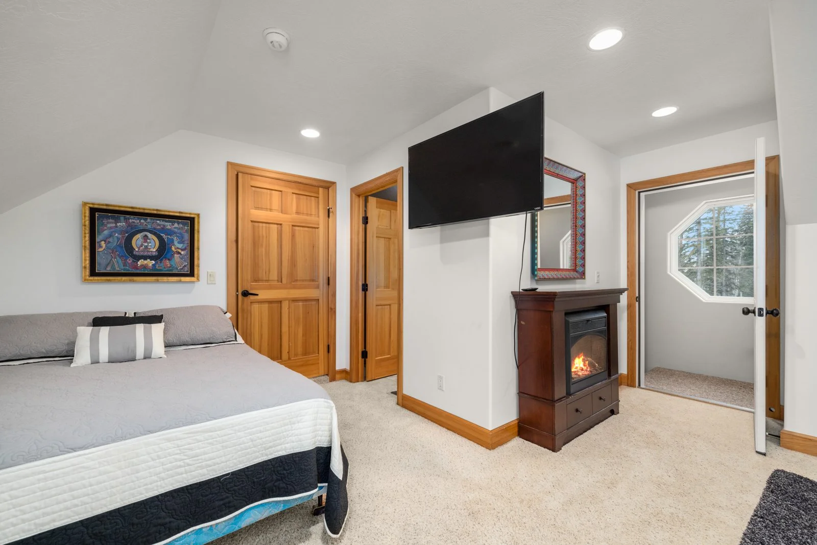 A cozy bedroom with a bed, wooden closet doors, a wall-mounted TV, a fireplace with a mirror above, and a window showing trees outside.
