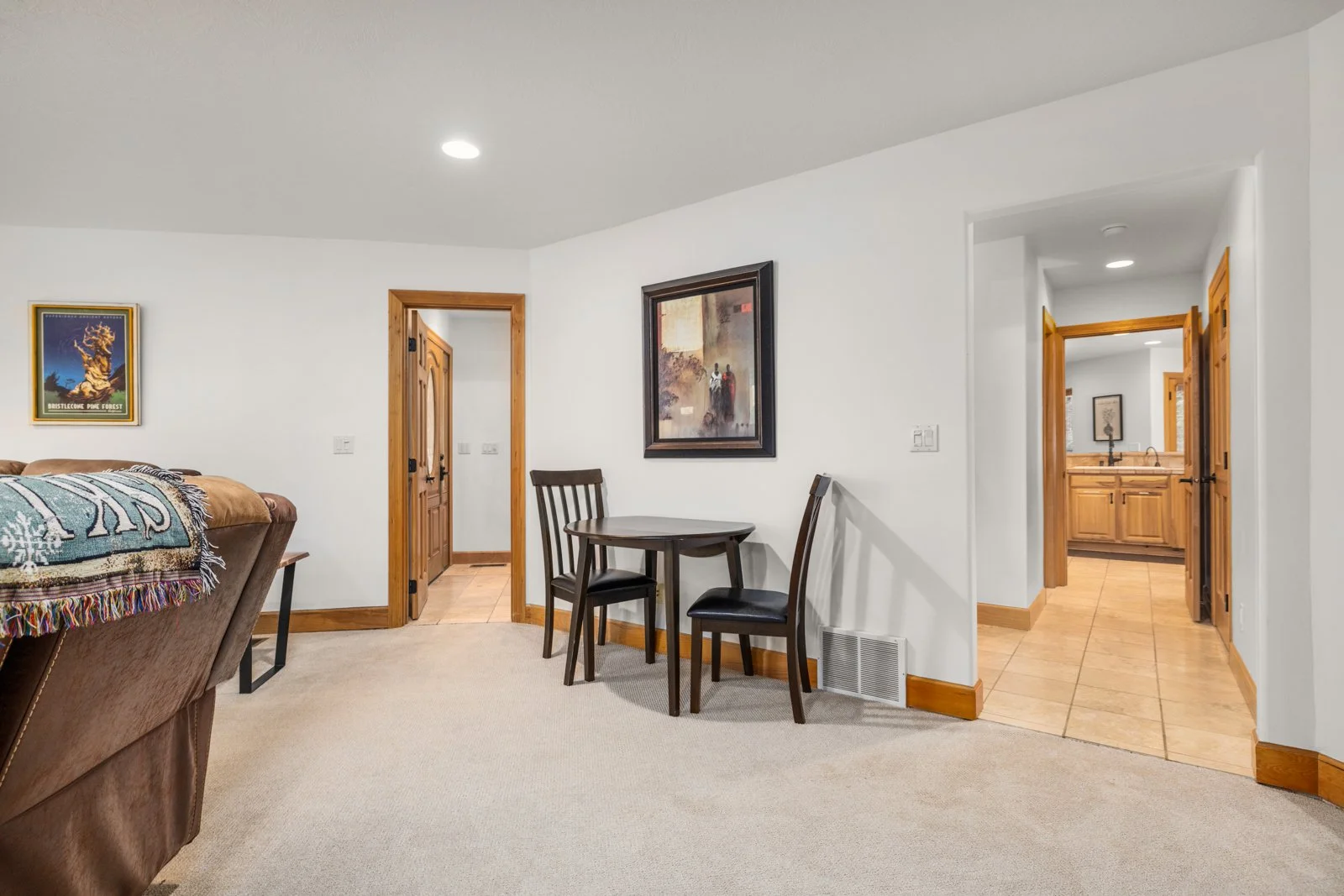 Living room with a brown sofa, a small round table with two chairs, framed artwork on white walls, and two doors leading to other rooms with wooden trim, one with tiled flooring.