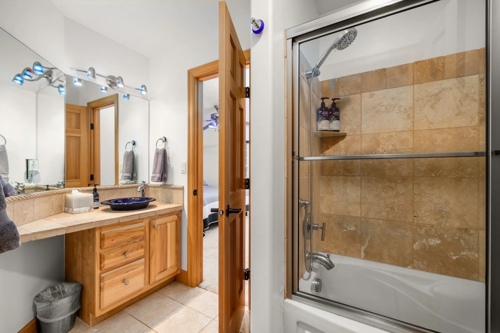 Bathroom with wooden vanity, large mirror, gray towels, and a shower with tan tiles and soap bottles.