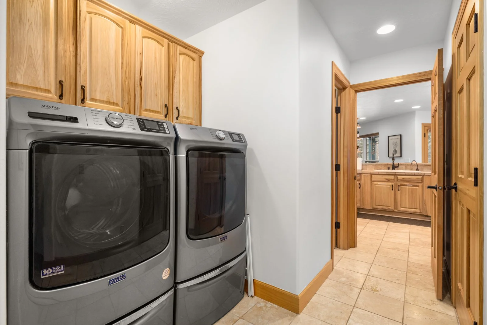Laundry room with a silver washer and dryer side by side, wooden cabinets above, beige tiled floor, and an open door leading to a bathroom with wooden cabinetry and a sink.