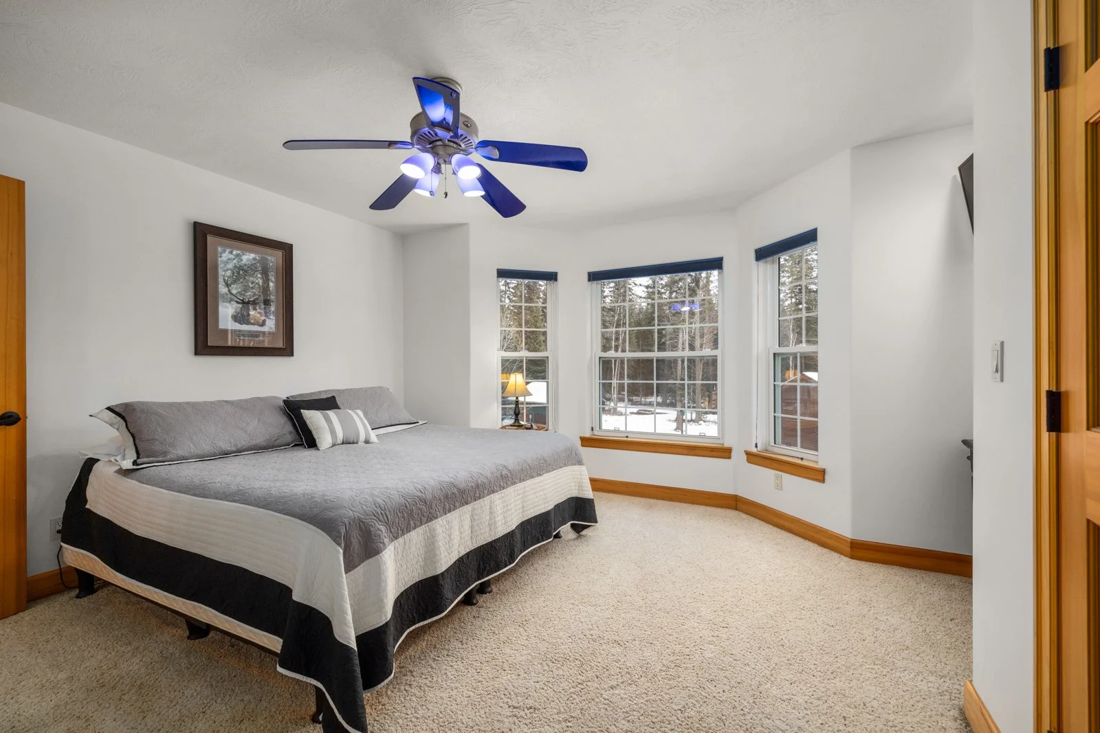 Bedroom with a large bed, a ceiling fan with lights, three windows with snow outside, and a small lamp on a side table.