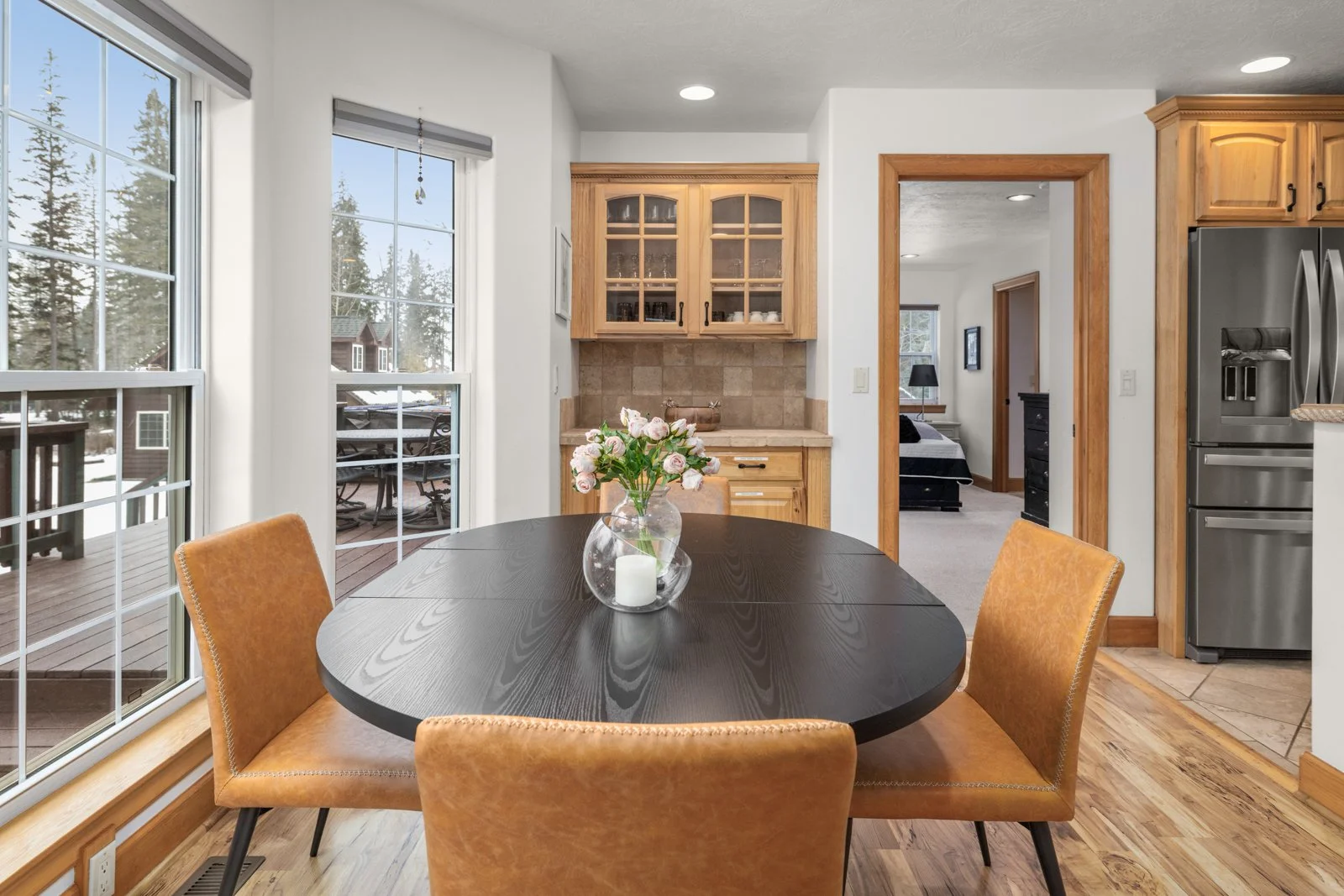 Kitchen and dining area with a round table, four tan chairs, a vase with flowers, large windows, a door leading outside, and a view into a bedroom.