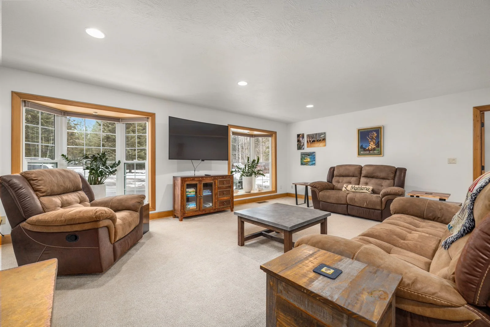 Living room with beige carpet, brown reclining sofas, a wooden coffee table, a television on a wooden stand, large windows showing snow outside, wall art, and potted plants.