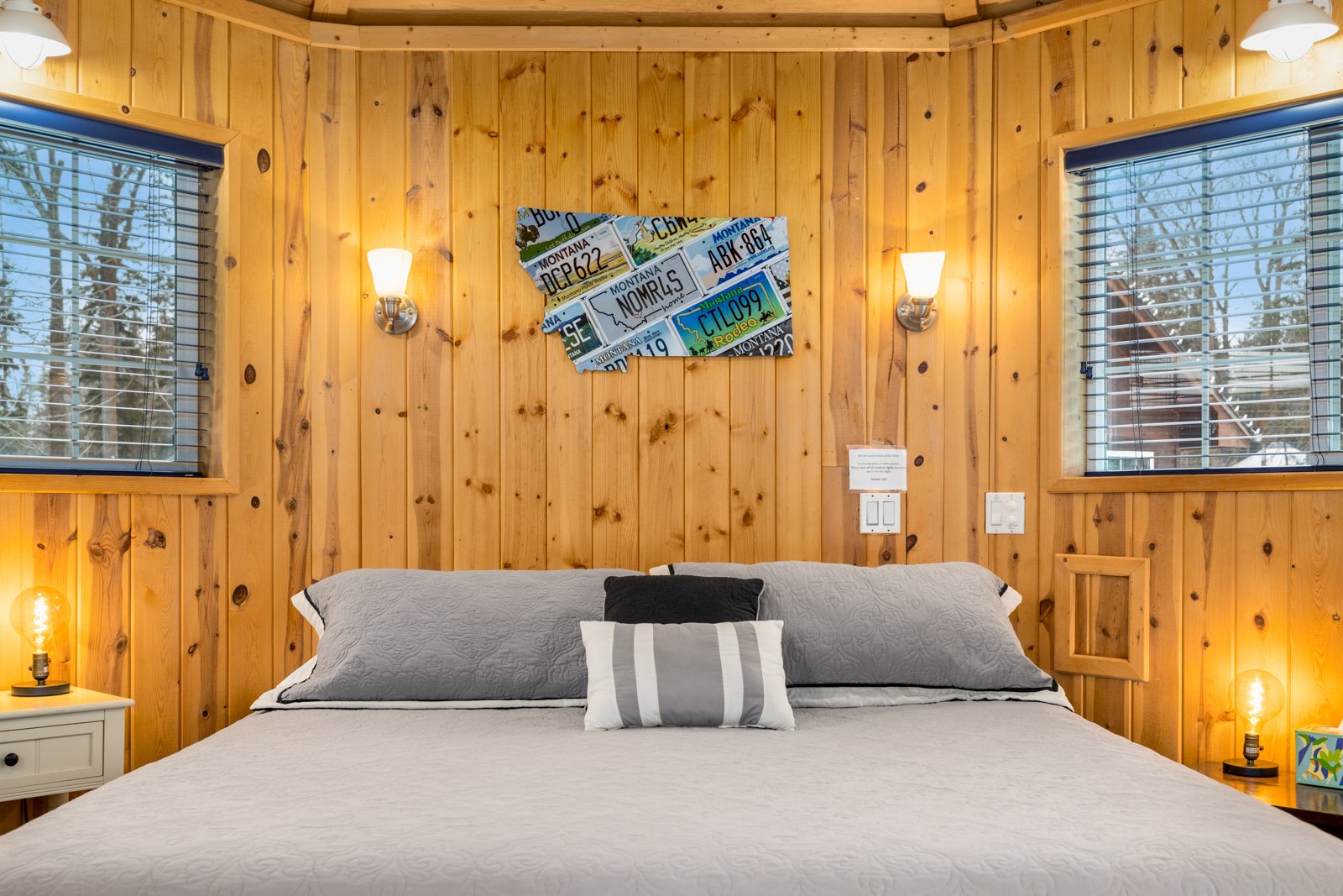 A bedroom with wooden panel walls, two windows with blinds, a bed with gray and black pillows, nightstands with lamps, and a wall art piece featuring various license plates.