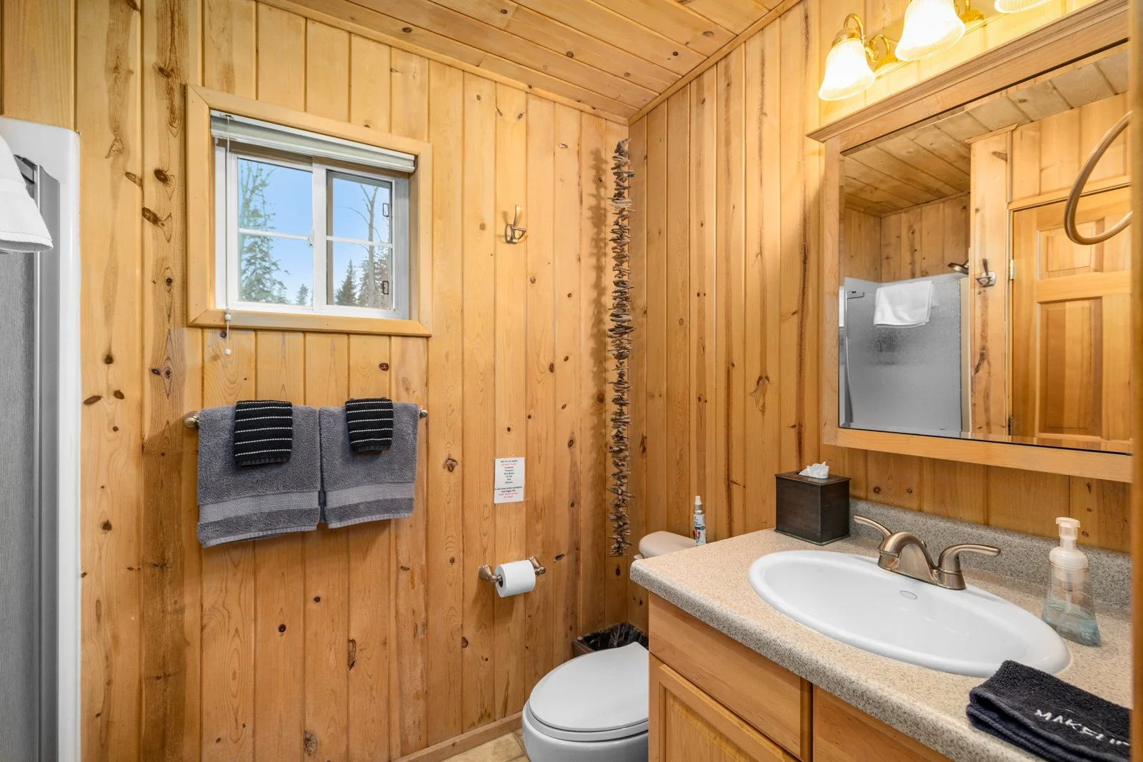 A rustic bathroom with wood-paneled walls, a small window, a toilet, a sink with a beige countertop, a mirror, and warm lighting.
