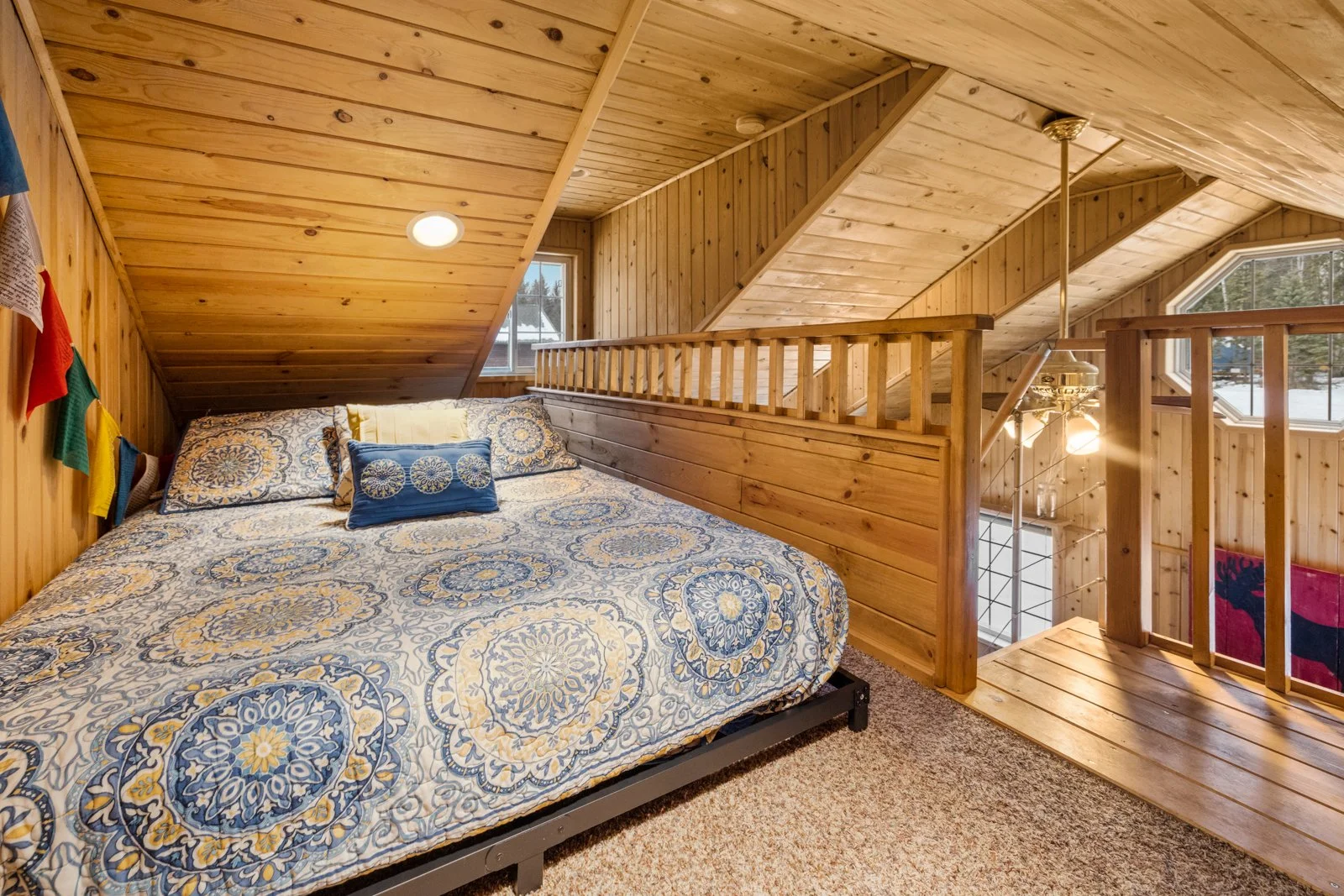 Wood-paneled attic bedroom with a bed featuring blue, yellow, and white patterned bedding, and a small decorative pillow. There are small windows and a wooden railing around the partial upper level.