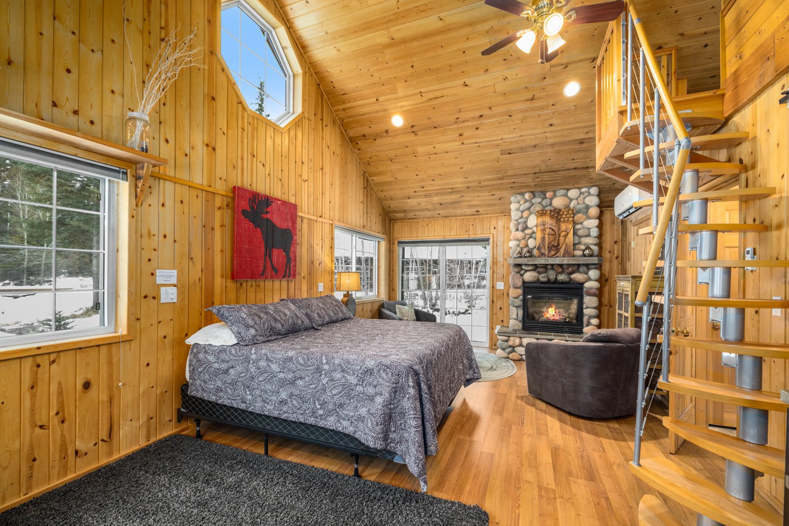 Interior of a cozy cabin bedroom with wooden walls and ceiling, a stone fireplace with a lit fire, a bed with patterned bedding, a lounge chair near the fireplace, and a spiral staircase leading upward.