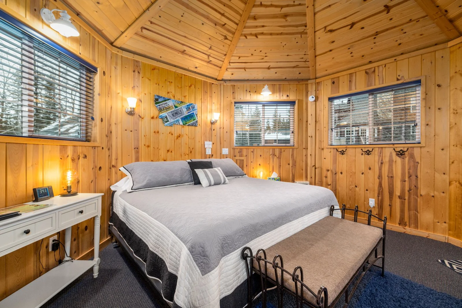 A cozy bedroom with wood-paneled walls and ceiling, featuring a large bed with gray and black bedding, a white nightstand with a lamp and clock, and three windows with blinds showing a winter scene outside.