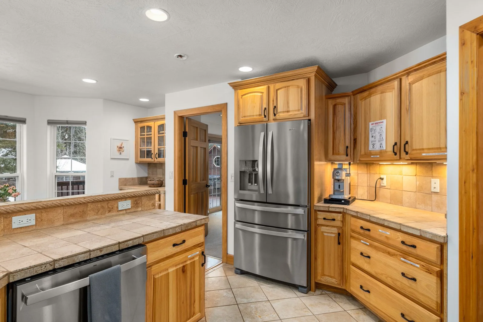 Kitchen with wooden cabinets, stainless steel refrigerator, tiled countertops, and a coffee machine on the counter.