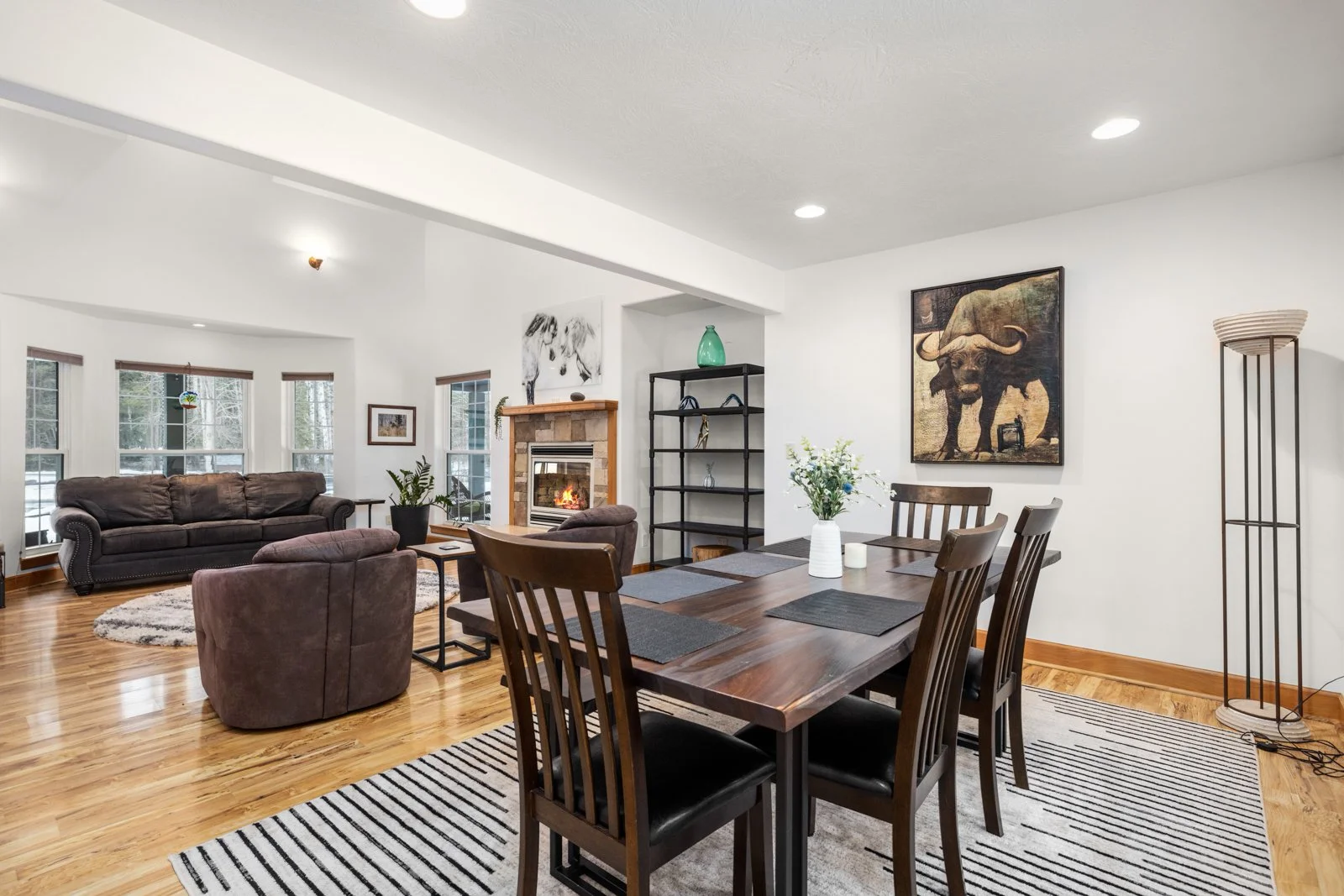 Open-concept living and dining room with white walls, hardwood floors, and multiple windows. There is a dining table with six chairs, a fireplace with a stone surround, a dark sofa, a matching armchair, a black bookshelf, wall art, a vase with flower