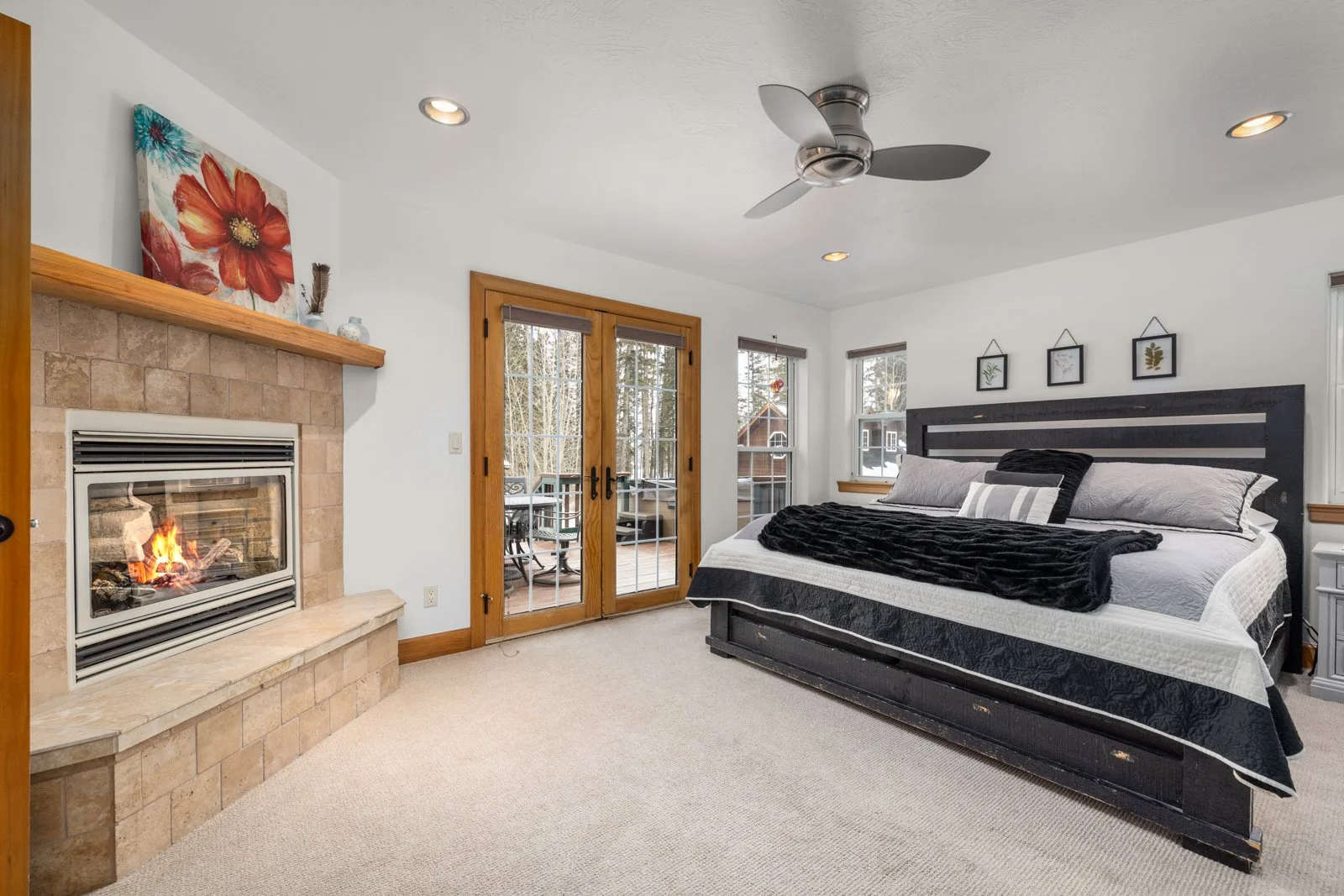 A bedroom with a large bed, black headboard, and gray bedding, next to a fireplace with a mantel, outside a set of glass doors leading to a patio, with multiple windows allowing natural light, hardwood trim, ceiling fan, and framed artwork on the wal