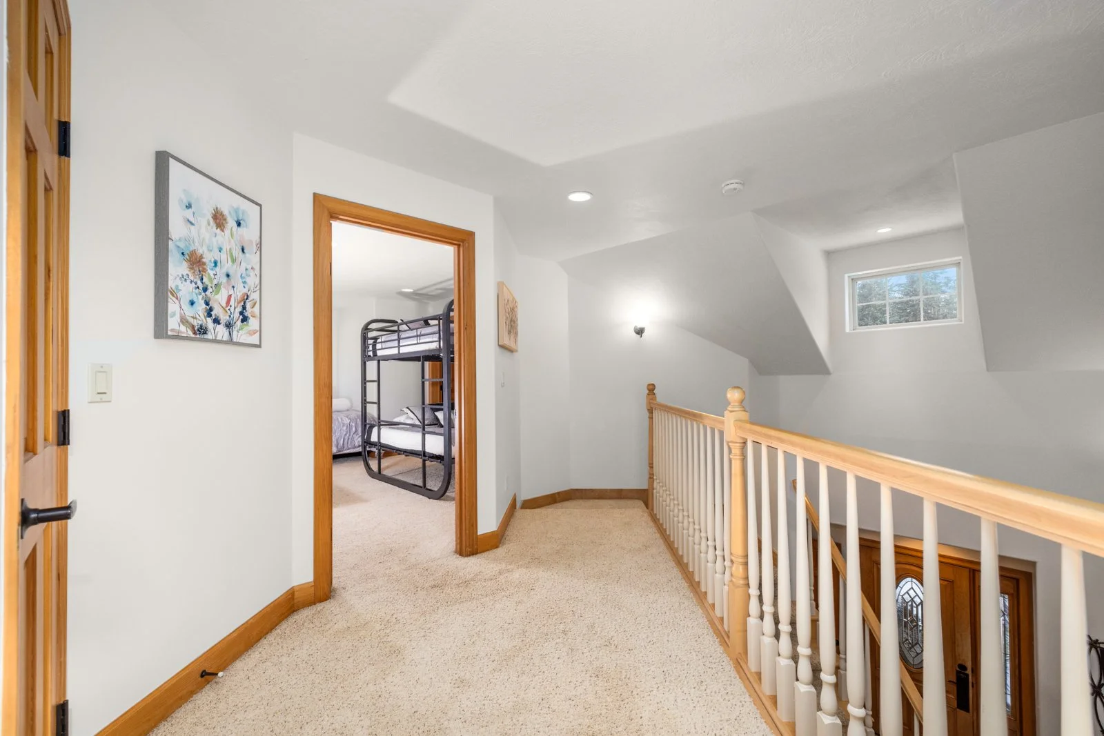 Upper-floor hallway with beige carpet, wooden trim, a stair railing, and an open doorway to a bedroom with bunk beds, window, and walls decorated with a colorful floral painting.