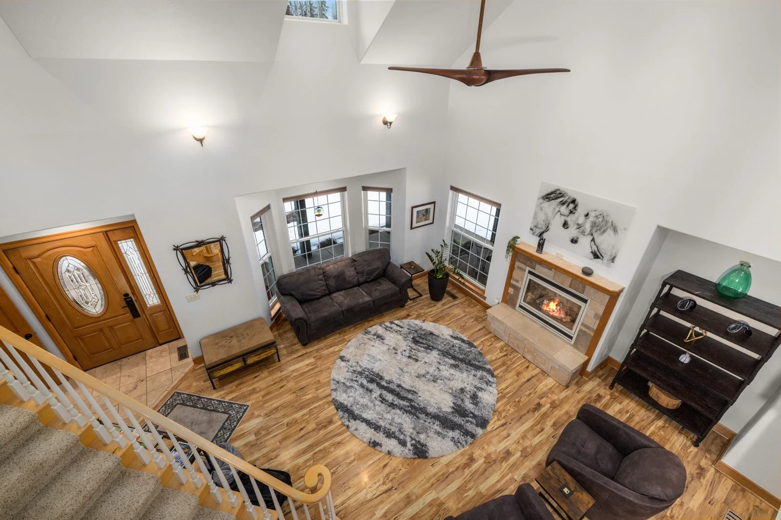 Living room viewed from staircase, featuring flooring, a round patterned rug, dark sofas, a stone fireplace with a wooden mantle, a large black and white horse artwork above the fireplace, a tall green vase, a black shelving unit with decorative item