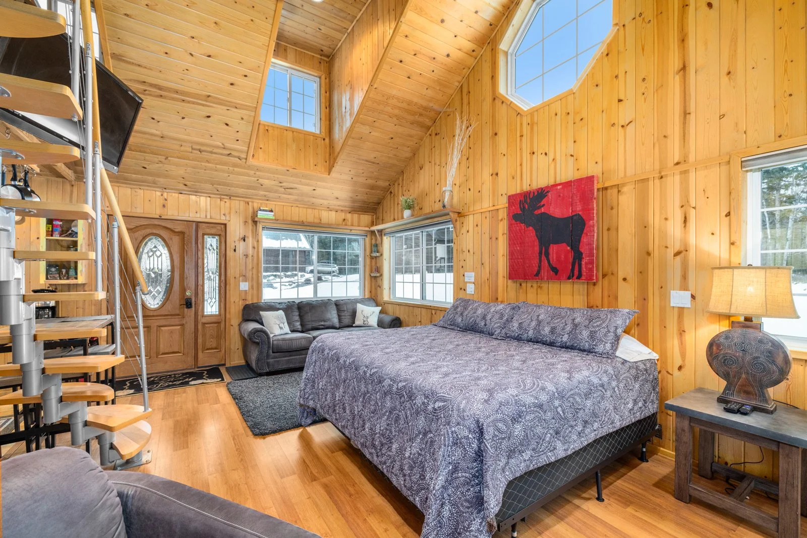 Interior view of a cozy wooden cabin living room with a queen-sized bed, a gray sofa, a small table with a lamp, and a colorful moose wall art.