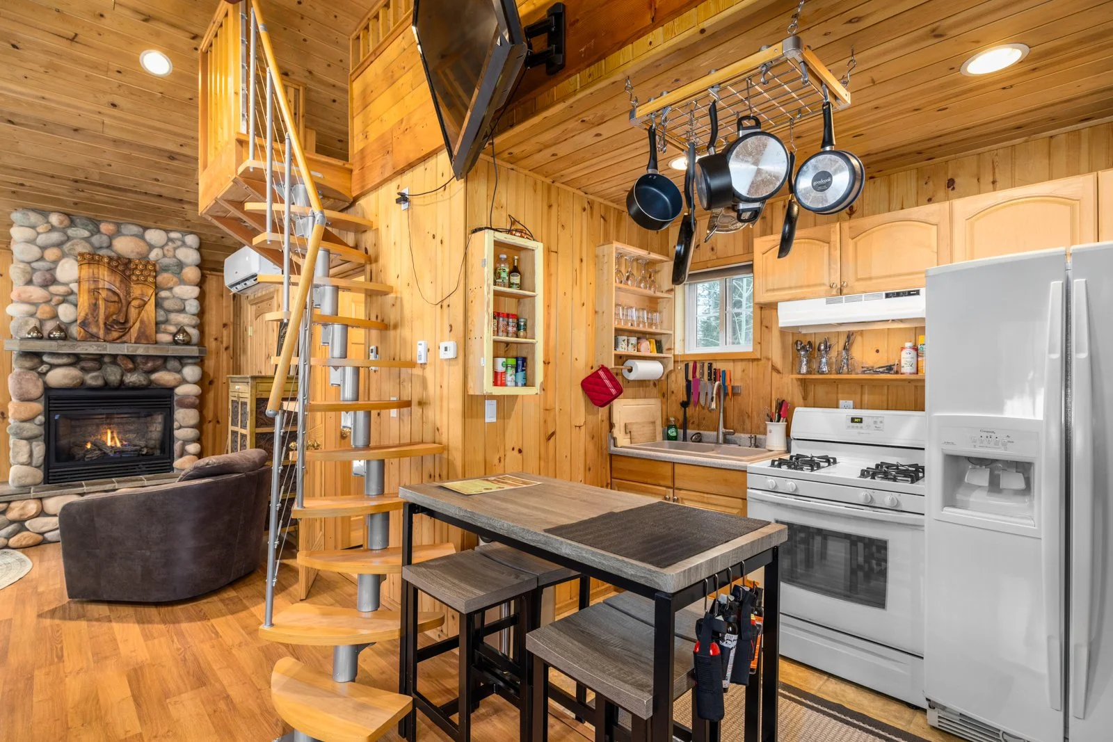 A cozy kitchen with wooden walls and ceiling, a gas stove, a refrigerator, and a small sink. A pot rack hangs above the kitchen area. Spiral staircase leads to a loft. Fireplace with a stone surround is in the background, along with a brown armchair 