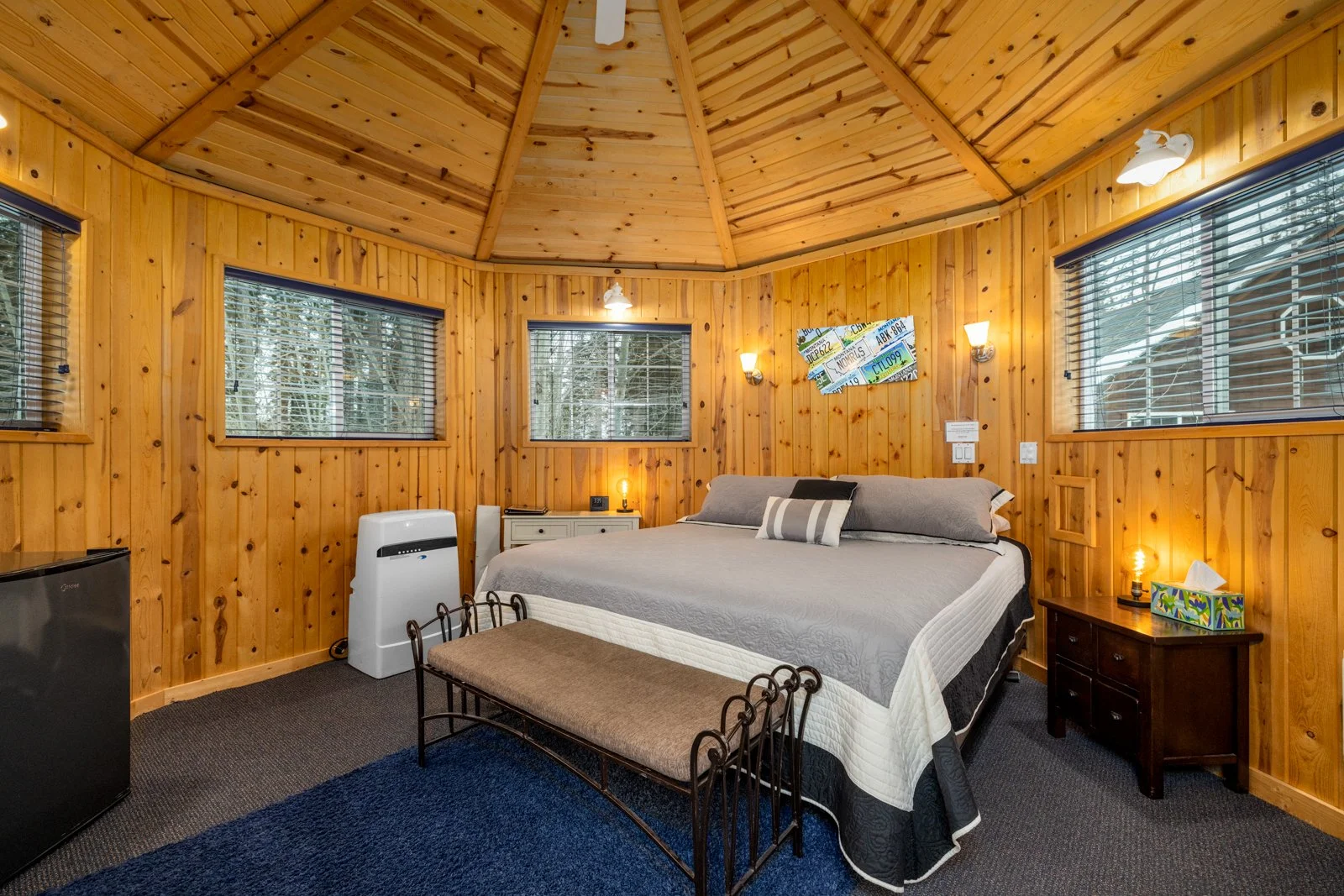 Bedroom with wood-paneled walls and ceiling, a large bed with gray and white bedding, a dark wooden side table with a tissue box, small lamp and a framed photograph, three windows with blinds, a small heater or air purifier, a bench at the foot of th