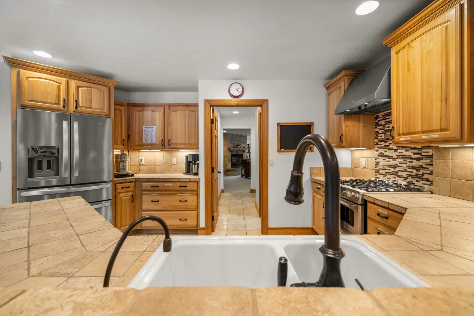 Kitchen with wooden cabinets, stainless steel refrigerator, black faucet, tiled countertops, gas stove, and hallway leading to a living room with fireplace.