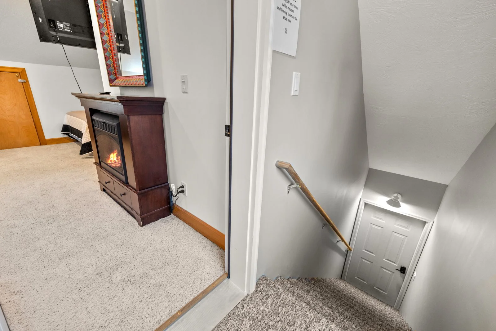 A split view of an interior space showing a room with a fireplace and television on the left and a staircase with a wooden handrail leading down to a closed door on the right.